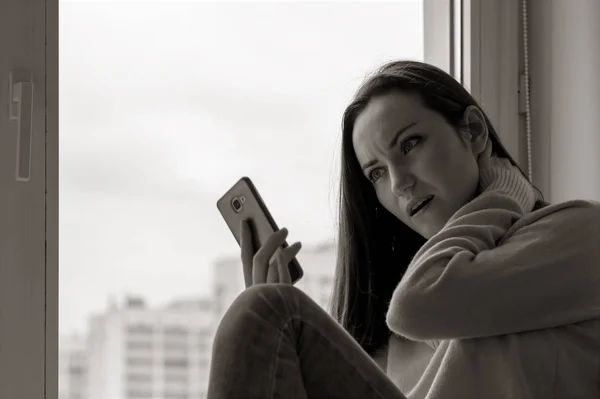 Een jonge vrouw is depressief op een vensterbank in de buurt van het venster van het huis, close-up, zwart-wit. — Stockfoto