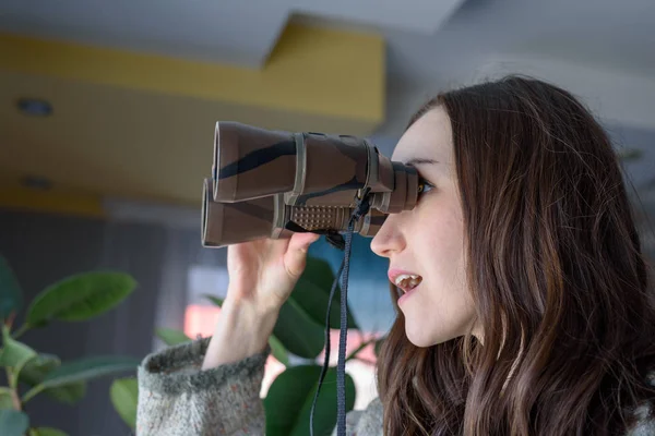 Young woman spying out the window looking through binoculars — Stock Photo, Image