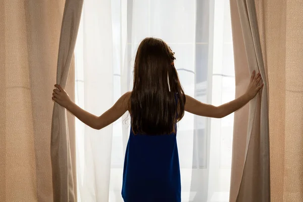 Het meisje opent de gordijnen bij het grote raam in het huis — Stockfoto