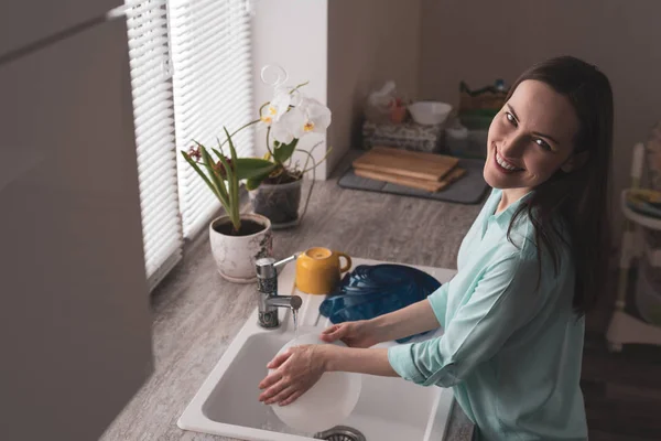 Mujer joven sonriente lavando platos en la cocina frente a una ventana con flores en macetas en la suave luz de la mañana, tonificado ángulo de escena auténtica desde la parte superior y desde el lado —  Fotos de Stock