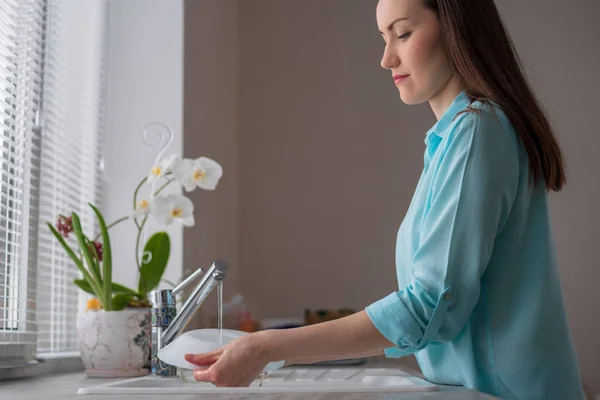 La chica en una blusa turquesa lava un plato blanco en el fregadero delante de la ventana con persianas —  Fotos de Stock