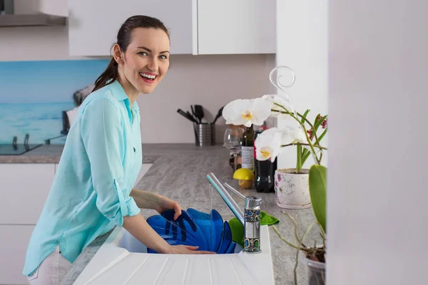 Retrato de una mujer sonriente en la camisa de color turquesa delante de la ventana en la cocina, enjuaga un plato azul — Foto de Stock