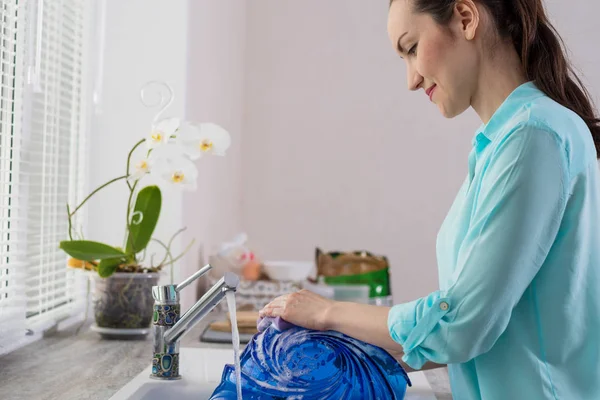 Retrato enmarcado de una ama de casa frente a una ventana en la cocina, lavando un plato azul bajo el agua corriente — Foto de Stock