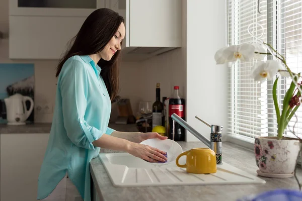 Una mujer con camisa turquesa lava los platos bajo un grifo en la cocina — Foto de Stock