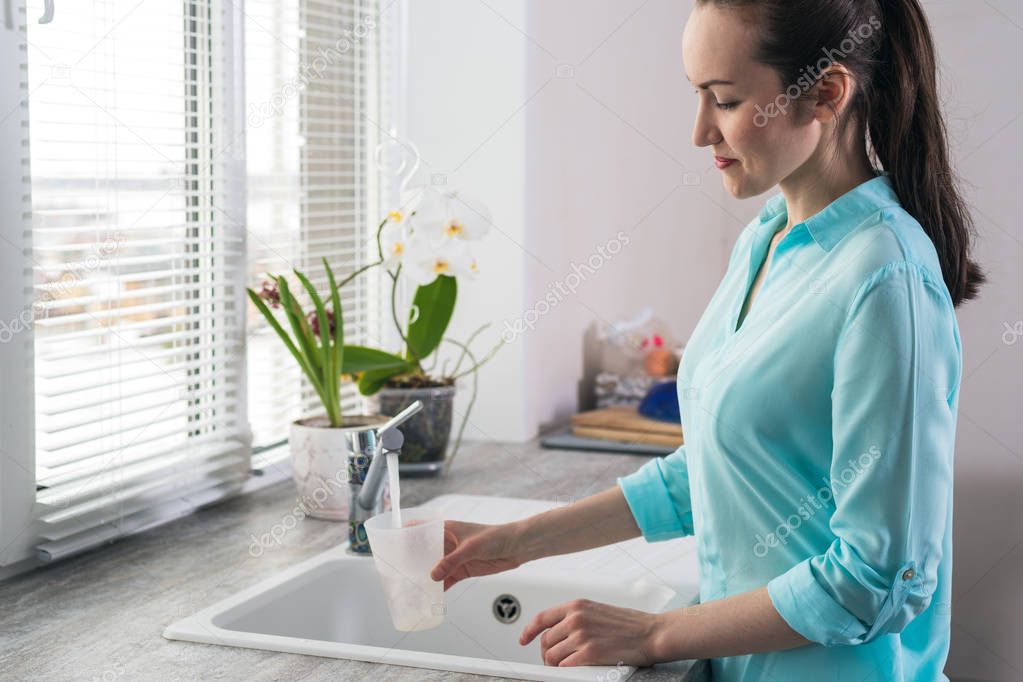 Portrait of a young woman gaining a glass of clean tap water in the kitchen in front of the window