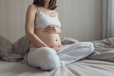 Hamile kadın elleri karnında, beyaz bluz ve mavi pantolonla yatakta oturuyor.