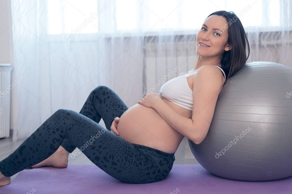 portrait of pregnant woman in white top and gray leggings sitting on gym Mat with gym ball