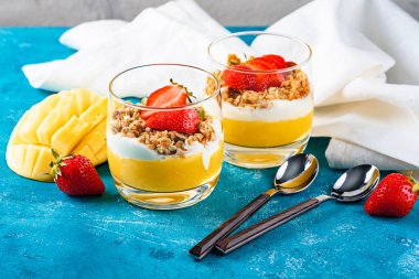 Mangolu ve çilekli iki bardak müsli ve kremalı tatlı veya turkuaz arka planda tatlı kaşıklarıyla ricotta.