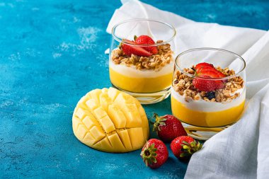 iki bardak mango ve müsli ve kremalı çilekli tatlı ya da turkuaz arka planda beyaz pamuklu kumaşlı ricotta.