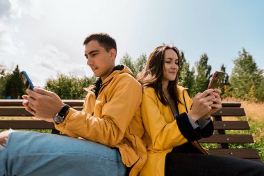Sonbahar Parkı 'ndaki bankta oturan sarı ceketli gülümseyen genç çift telefonları ve akıllı telefonları kullanıyor. Milenyum kuşakları, cihazlardan ayrılmaz.