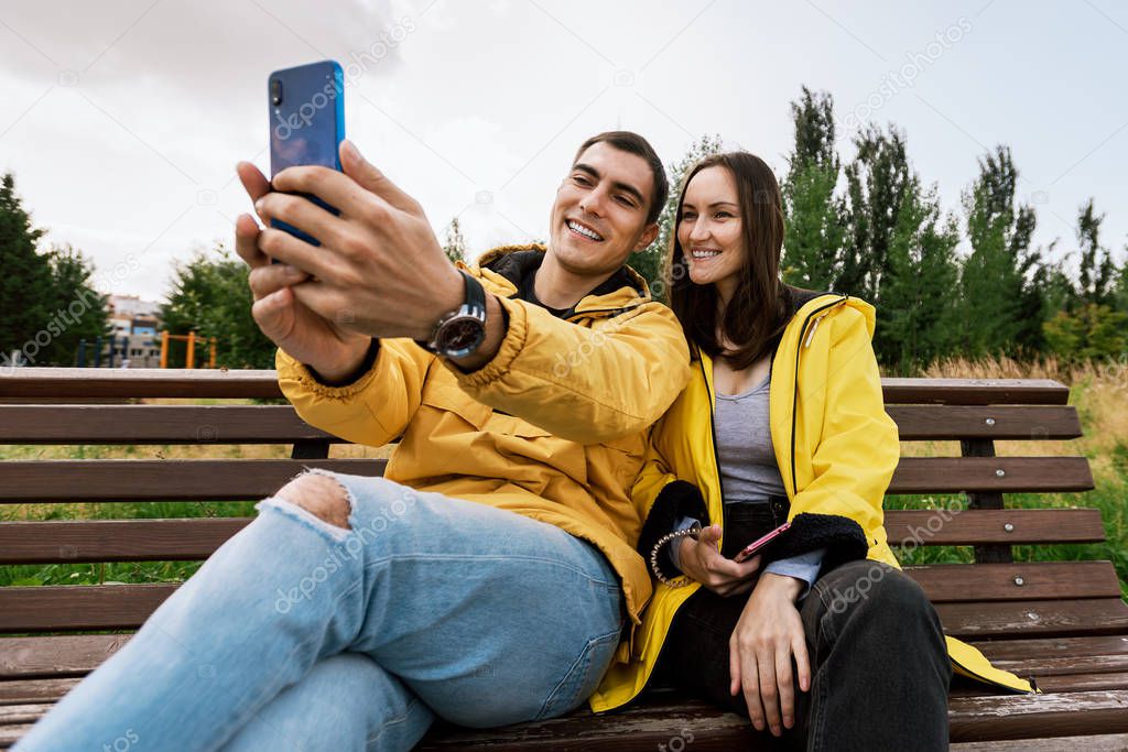 smiling young couple in yellow jackets take a selfie, photo on a smartphone, phone, sitting on a bench in the autumn Park. Millennials, inseparable from the gadget, device