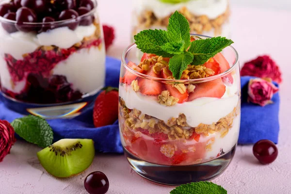 Trifles with cherries, strawberries, whipped cream and granola in glass on blue napkin on light pink background — Stock Photo, Image