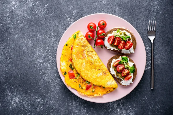 Sağlıklı bir omlet kahvaltısı, krem peynirli kepekli tost, pesto ve vişneli domatesler pembe tabakta, siyah taşlı arka planda uzay kopyası..