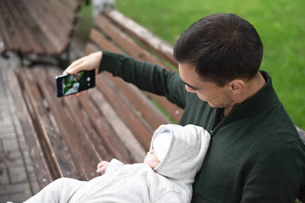 Babası yeşil kazaklı, kollarında bebekle bankta oturmuş selfie çekiyor. — Stok fotoğraf