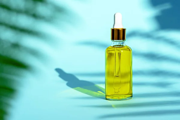 Dropper glass bottle with yellow cosmetic oil on blue background and leaf shadow — 스톡 사진