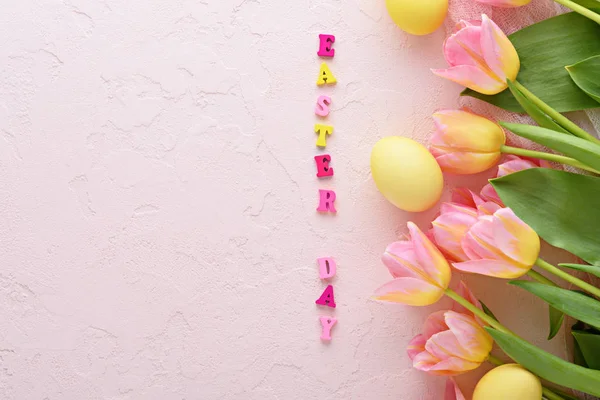 top view yellow pastel Easter eggs with pink tulips on pink background and easter day lined with multicolored letters