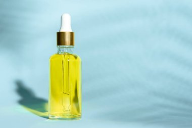 dropper glass bottle with pipette and yellow oil on blue background with leaf shadow and copy space
