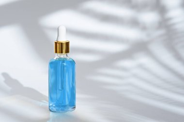 trendy dropper glass bottle with pipette and blue liquid on white background with leaf shadow, copy space