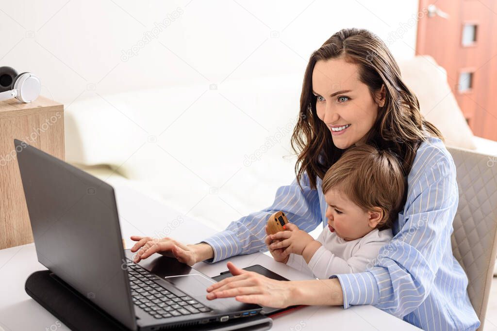 young smiling brunette mother in blue shirt working on laptop with her baby at home, working on maternity leave