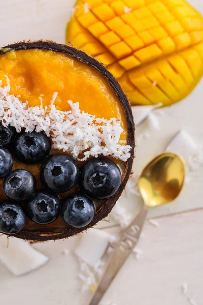 vertical smoothie bowl with mango mousse, coconut flakes, blueberries in coconut bowl with dessert spoon on white wooden background, mango ice cream concept, eco dessert