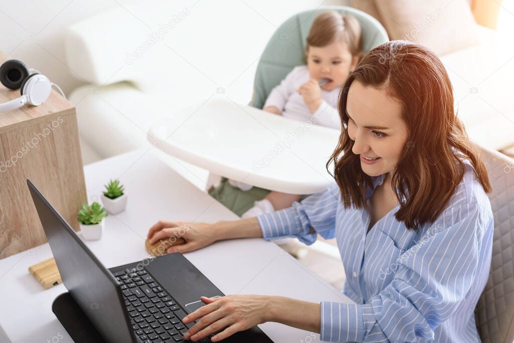 smiling caucasian woman in blue shirt is working at laptop, child sitting next to her, mother on maternity leave, freelancer, remote work at home concept