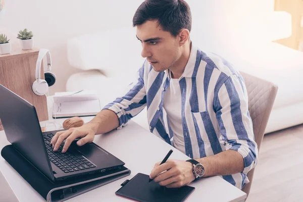 dark-haired man Illustrator in blue shirt draws on graphic tablet, freelancer, remote work on laptop at home, graphic designer concept