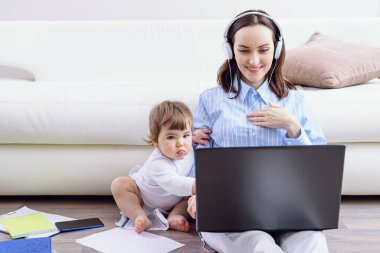 woman with headphones looks at laptop screen, child is sitting next to her, home-learning concept, remote work at home clipart