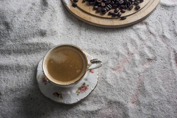 A cup of coffee, and grains of coffee on white sand on a blue background