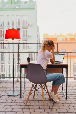 Genç yorgun kadın terasta dizüstü bilgisayar kullanıyor.