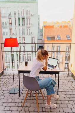Genç yorgun kadın terasta dizüstü bilgisayar kullanıyor.