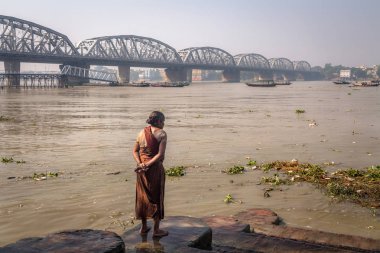 Old woman watches high tide water flow in the Ganges river at Dakshineshwar. The Bally bridge (Vivekananda Setu) at the backdrop. clipart