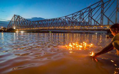 Günbatımında Ganj nehri üzerinde tarihi Howrah köprüsü. Kolkata Hindistan 'daki bir ayinin bir parçası olarak nehirde kil diya lambaları taşıyan yaşlı bir kadın görülüyor.