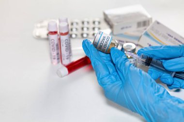 Doktor ellerinde şırınga ile Covid-19 tedavisinde kullanılan bir aşı şişesi var. Arka planda tıbbi ilaçlar ve kan örnekleri var.