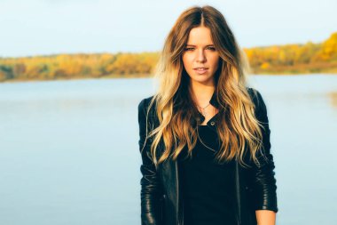 Genç güzel sarışın bir kadınla sonbahar Park mükemmel dişleri olan koyu deri ceket gülümsemek günbatımı sırasında gölde.