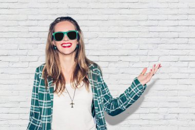 Genç güzel yeşil kareli ceket ve güneş gözlüğü mevcut kol jest kentsel mutlu hippi kız yapmak, gülmek ve geniş ile mükemmel dişler gülümseme. Beyaz tuğla duvar arka plan.