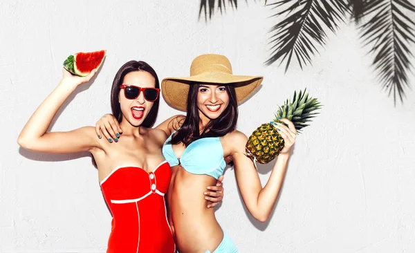 Відпустка. Літні поїздки. Дві красиві сексуальні брюнетки-близнюки відкриті з кавуном і ананасами в бікіні на білому тлі стіни з тіні пальми весело посміхаються на пляжі — стокове фото