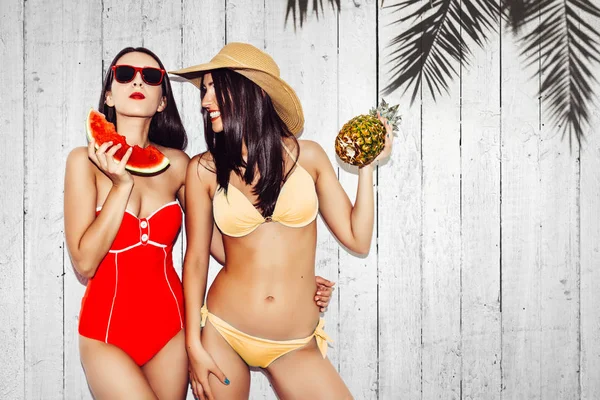Відпустка. Літні поїздки. Дві красиві сексуальні брюнетки на відкритому повітрі з кавуном і ананасами в бікіні весело посміхаються на пляжі. Дерев'яний фон з простором для копіювання пальми для тексту — стокове фото