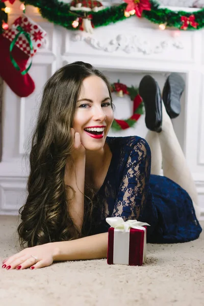 Christmas Woman. Beauty Model Girl with Fireplace on Background. Christmas Gift in Hand. Open Mouth Beautiful Teeth Smile. True Emotions. Red Lips and Manicure. Beautiful Holiday Makeup. Not isolated