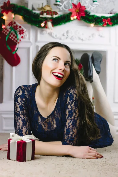 Christmas Woman. Beauty Model Girl with Fireplace on Background. Christmas Gift in Hand. Open Mouth Beautiful Teeth Smile. True Emotions. Red Lips and Manicure. Beautiful Holiday Makeup. Not isolated