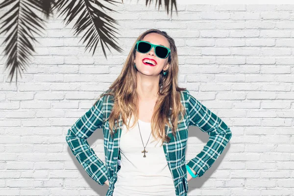 Genç güzel kentsel mutlu hipster bin yıllık kız yeşil kareli ceket ve eğlence, kahkaha ve mükemmel dişleri geniş gülümseme sahip güneş gözlüğü. Beyaz duvar palm arka plan. Yakın çekim. Sıcak renk.