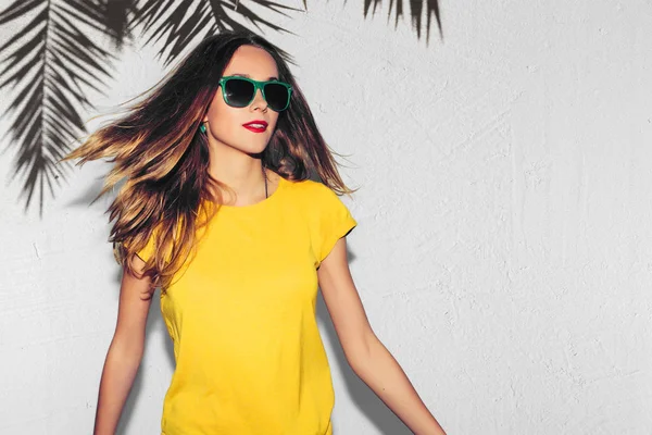 Hipster eller kalçalara palmiye ağacı arka plan üzerinde gülümseyen kız moda yaz portresi. Eğlenceli olumlu genç akıllı kadın gözlük ve mükemmel ile gülümseyen renkli sarı tişört.