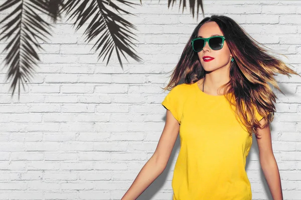 Hipster eller kalçalara palmiye ağacı arka plan üzerinde gülümseyen kız moda yaz portresi. Eğlenceli olumlu genç akıllı kadın gözlük ve mükemmel ile gülümseyen renkli sarı tişört.