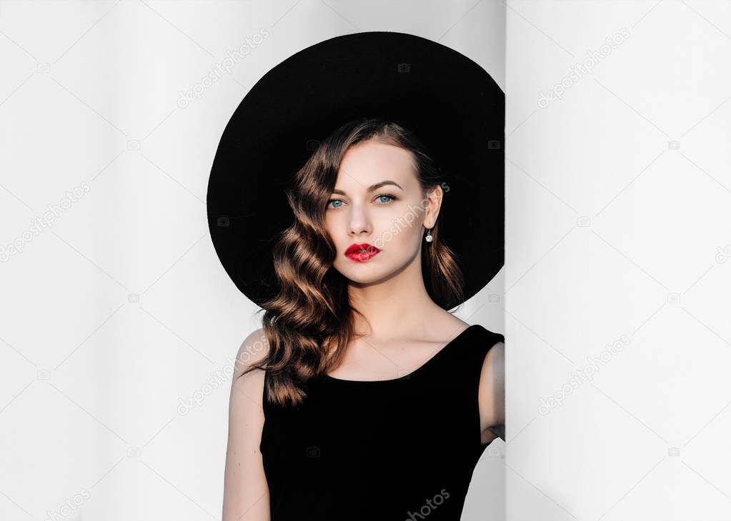 High fashion outdoor portrait of elegant woman in black hat and dress standing behind white pillar. Vintage-style portrait of a woman in black with red lips.