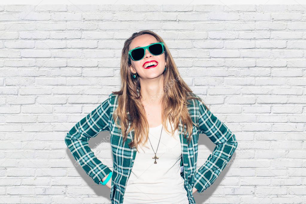 Young beautiful long-haired urban happy hipster millennial girl in green checkered jacket and sunglasses having fun, laugh and wide smile with perfect teeth. White brick wall background. Close up. Warm color.