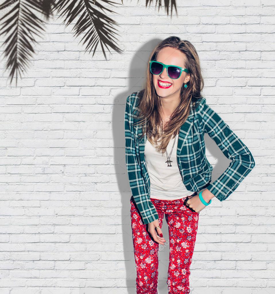 Young beautiful urban happy hipster millennial girl in green checkered jacket and sunglasses having fun, laugh and wide smile with perfect teeth. White wall palm background. Close up. Warm color.