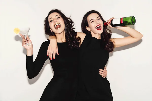 Duas mulheres bonitas em vestido de moda noite preto posando isolado em um fundo cinza branco. Menina morena bonita amigos gêmeos se divertindo bebendo coquetéis. Cantando e dançando. Garrafa de vinho . — Fotografia de Stock