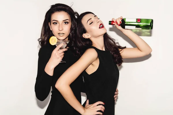 Duas mulheres bonitas em vestido de moda noite preto posando isolado em um fundo cinza branco. Menina morena bonita amigos gêmeos se divertindo bebendo coquetéis. Cantando e dançando. Garrafa de vinho . — Fotografia de Stock