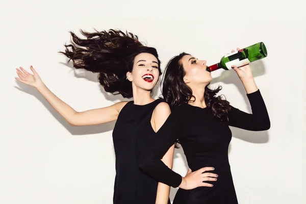 Duas mulheres bonitas em vestido de moda noite preto posando isolado em um fundo cinza branco. Menina morena bonita amigos gêmeos se divertindo bebendo coquetéis. Cantando e dançando. Humor festivo. Para dentro — Fotografia de Stock