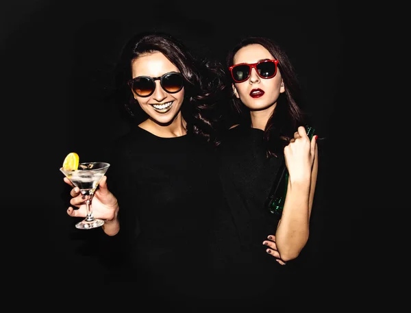 Două femei frumoase în rochie de modă de noapte neagră pozând izolate pe un fundal negru. Destul de bruneta fata prieteni gemeni având distractiv bea cocktail-uri. Cântând şi dansând. Sticlă de alcool . — Fotografie, imagine de stoc