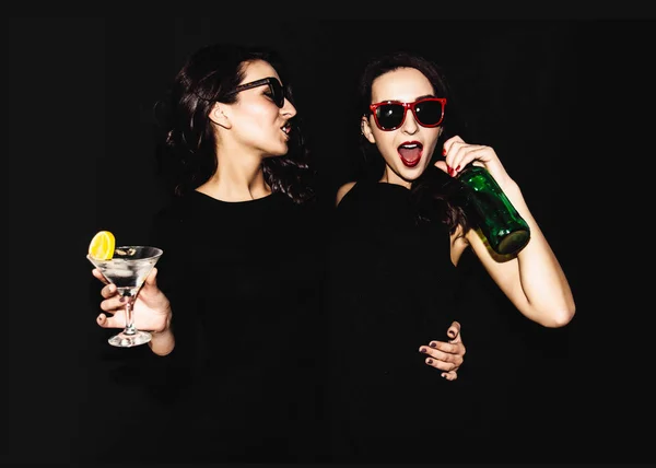 Duas mulheres bonitas em vestido de moda noite preta posando isolado em um fundo preto. Menina morena bonita amigos gêmeos se divertindo bebendo coquetéis. Cantando e dançando. Copo de garrafa de álcool . — Fotografia de Stock